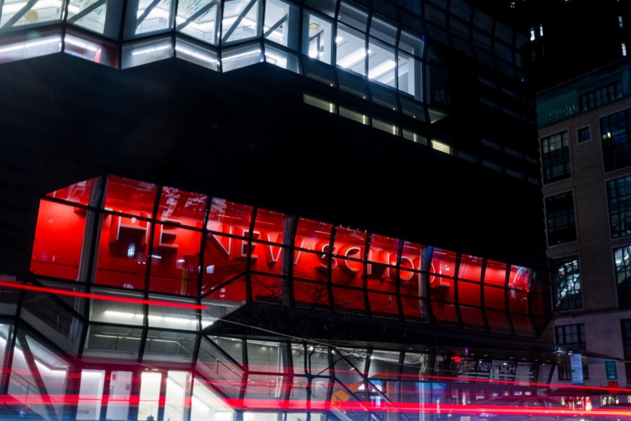 The facade of The New School's Fifth Avenue building at night. Letters behind large windows spell out the university's name and the lights of passing cars are blurred.