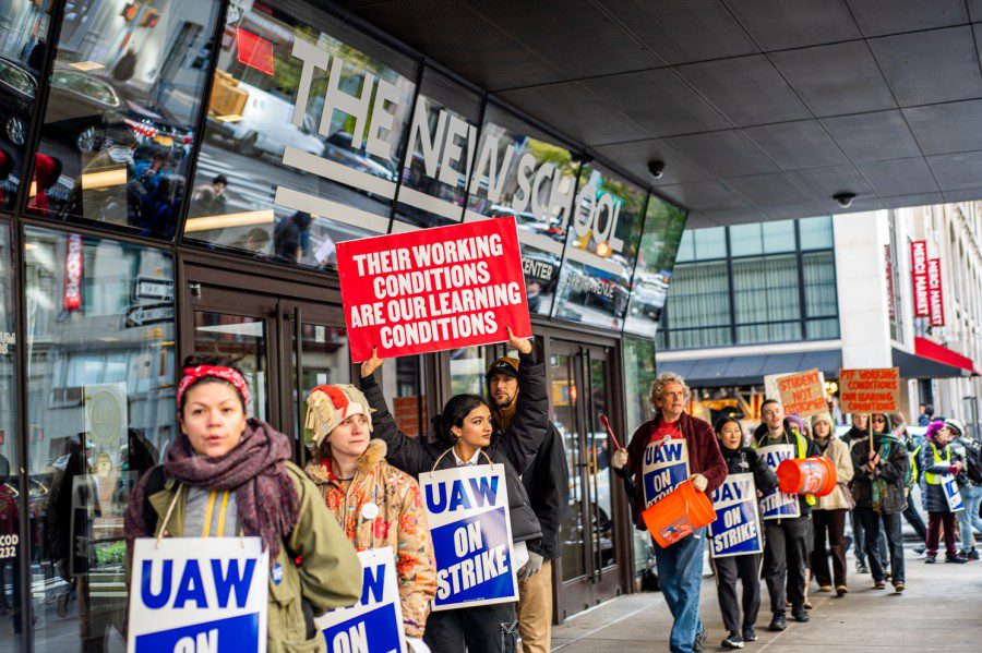 A group of part-time faculty members on strike in a picket line. They carry signs that read “U.A.W. on strike” and “Their working conditions are our learning conditions.” Behind them is the entrance to The New School’s main building.