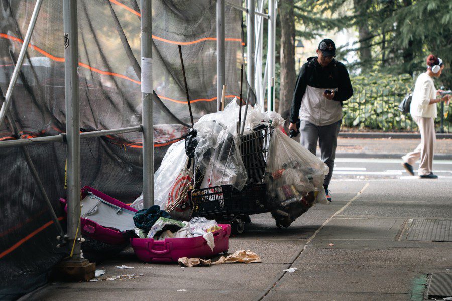 A cart wrapped around by plastic bags and an opened pink suitcase with clothes lays on a sidewalk under scaffolding.