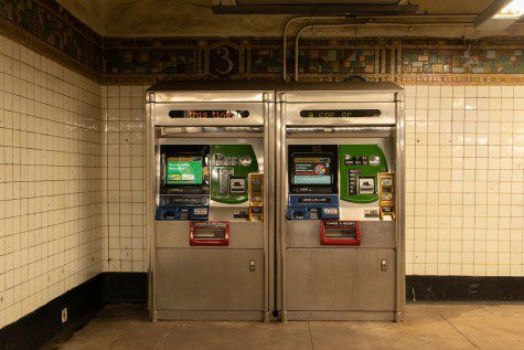 Two silver M.T.A. Metrocard vending machines with red, yellow, blue and green panels sit against a white-tiled wall.