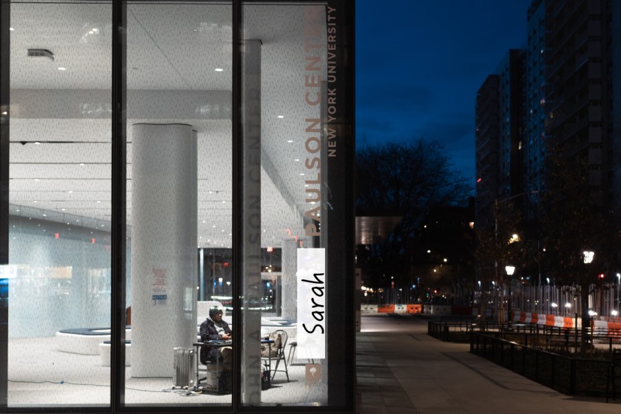 A corner of a building with text “Paulson Center New York University” on the glass in a vertical layout. Overlaying part of the original text is a block of handwritten text “Sarah.”