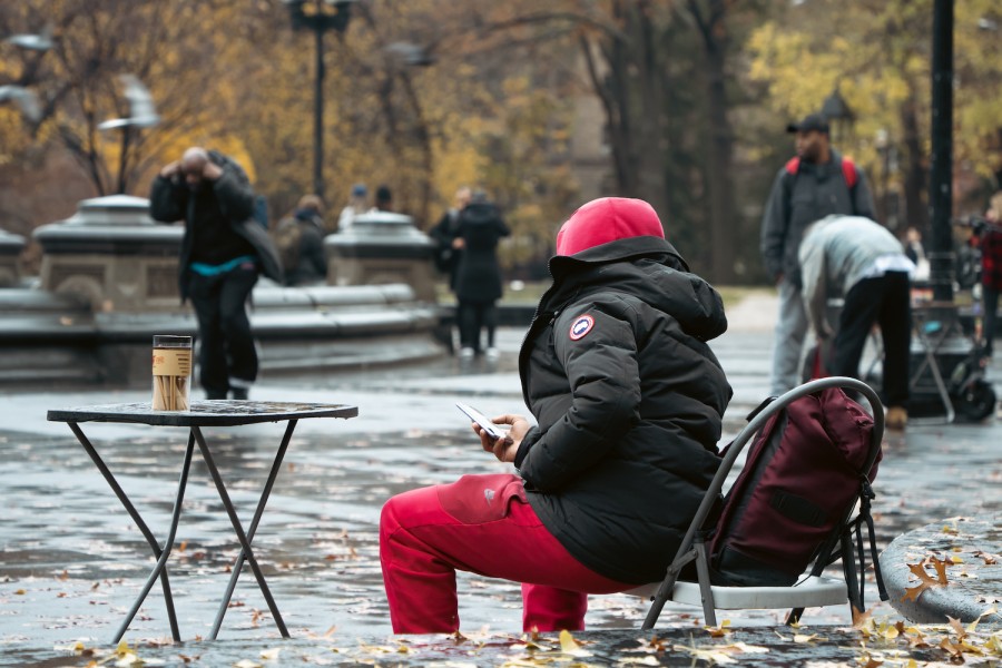 A+person+wearing+a+black+coat%2C+red+hoodie+and+red+pants+sits+on+a+stone+bench+in+a+park+with+a+small+table+in+front+of+them.+On+the+table+is+a+glass+jar+with+cannabis+joints+in+it.