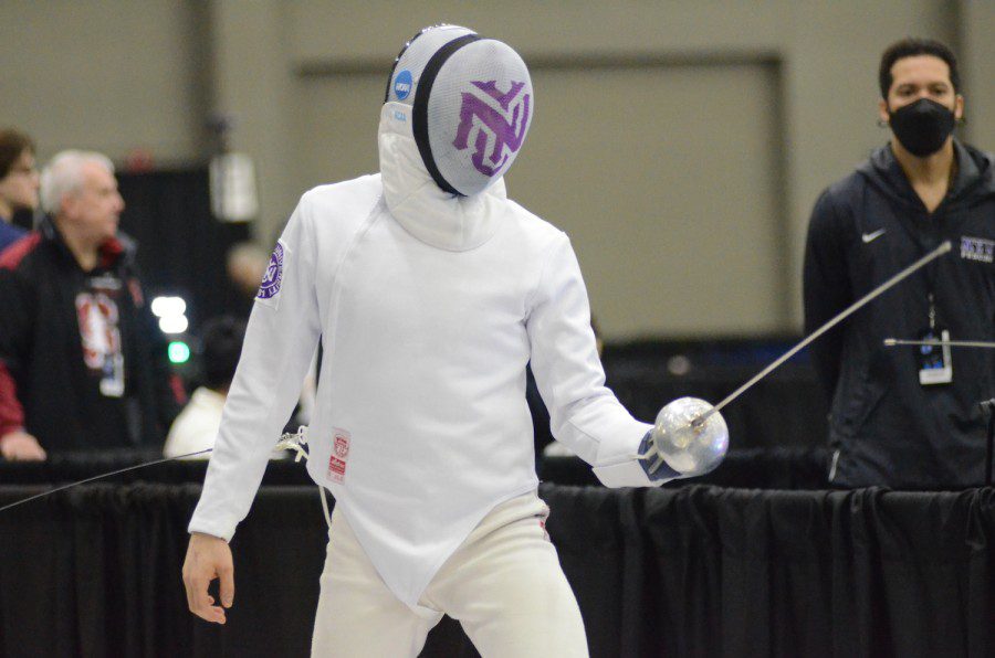 A fencer holding a silver foil dressed in white suit and a mask with a purple N.Y.U. logo printed on it.