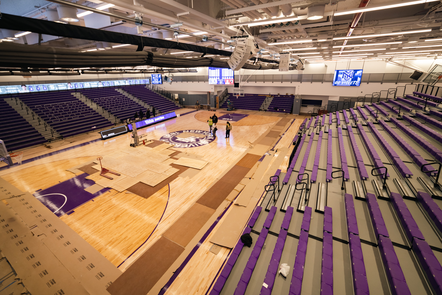 An indoor basketball court with white walls and two grandstands with purple seats on the two sides. The floor is partially covered with cardboard. Screens on the wall are displaying the logo of New York University’s athletic teams.