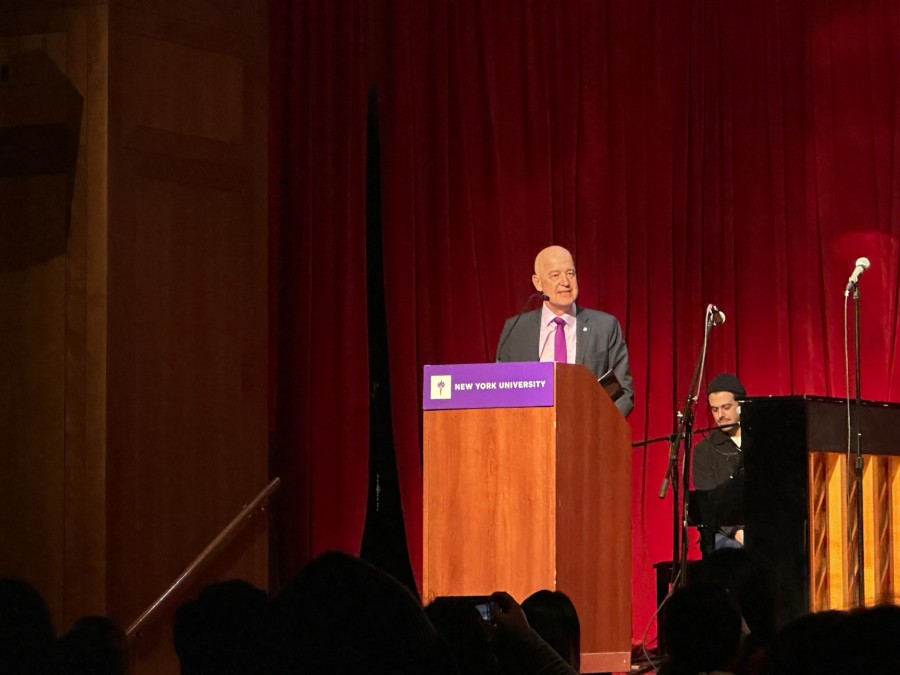 Andrew Hamilton, wearing a dark-colored suit with a red tie, stands behind a wooden N.Y.U. podium, on stage in an auditorium. Red curtains line the rear of the stage. An audio technician stands behind Hamilton.