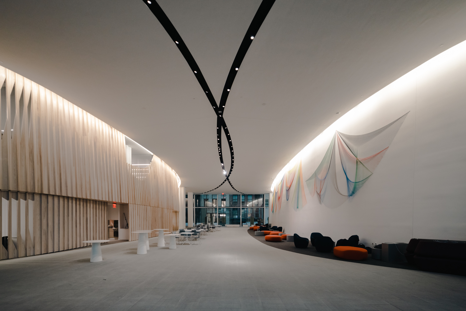 A large open lobby with two stripes of lights on the ceiling. There are white tables on the left side and red sofa chairs on the right side. On the wall to the right hangs an art installation with strands of colorful wires.