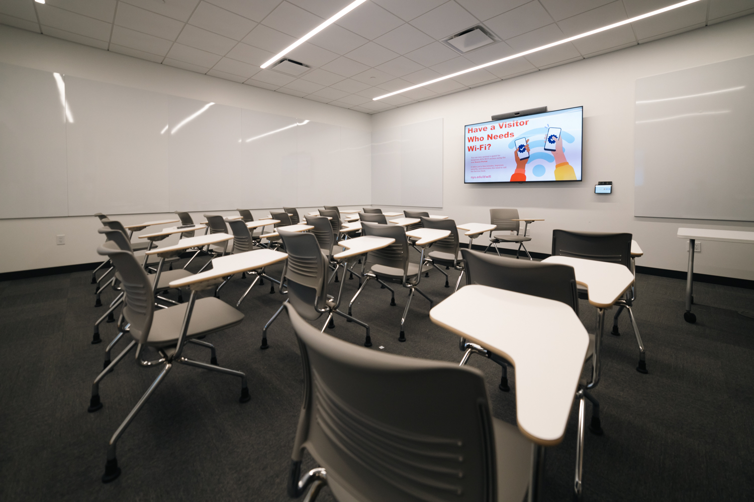 A classroom with a television hung on a white wall between two white boards. There are rows of gray chairs with small tables attached to them.