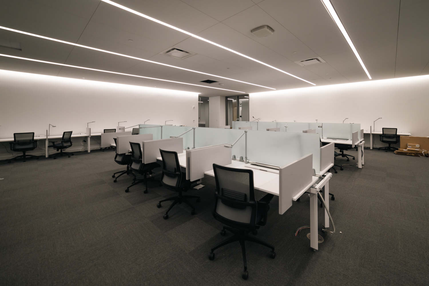 A quiet study room with white walls and several rows of white desks and black office chairs. The desks are separated from each other with boards made from glass and wood.