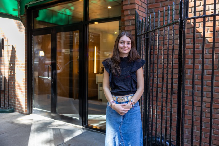 Alexa Donovan stands in front of 211 Thompson St., an apartment building in Greenwich Village. Alexa is looking at the camera and wearing a navy shirt and denim skirt.