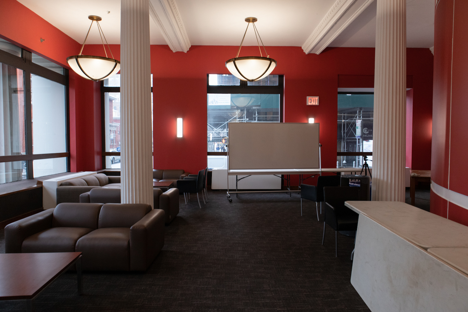 A student lounge on the first floor of the Silver Center. The room has bright, red walls lined with large windows, as well as couches, chairs, tables and a whiteboard inside. Two large lights hang from the ceiling.