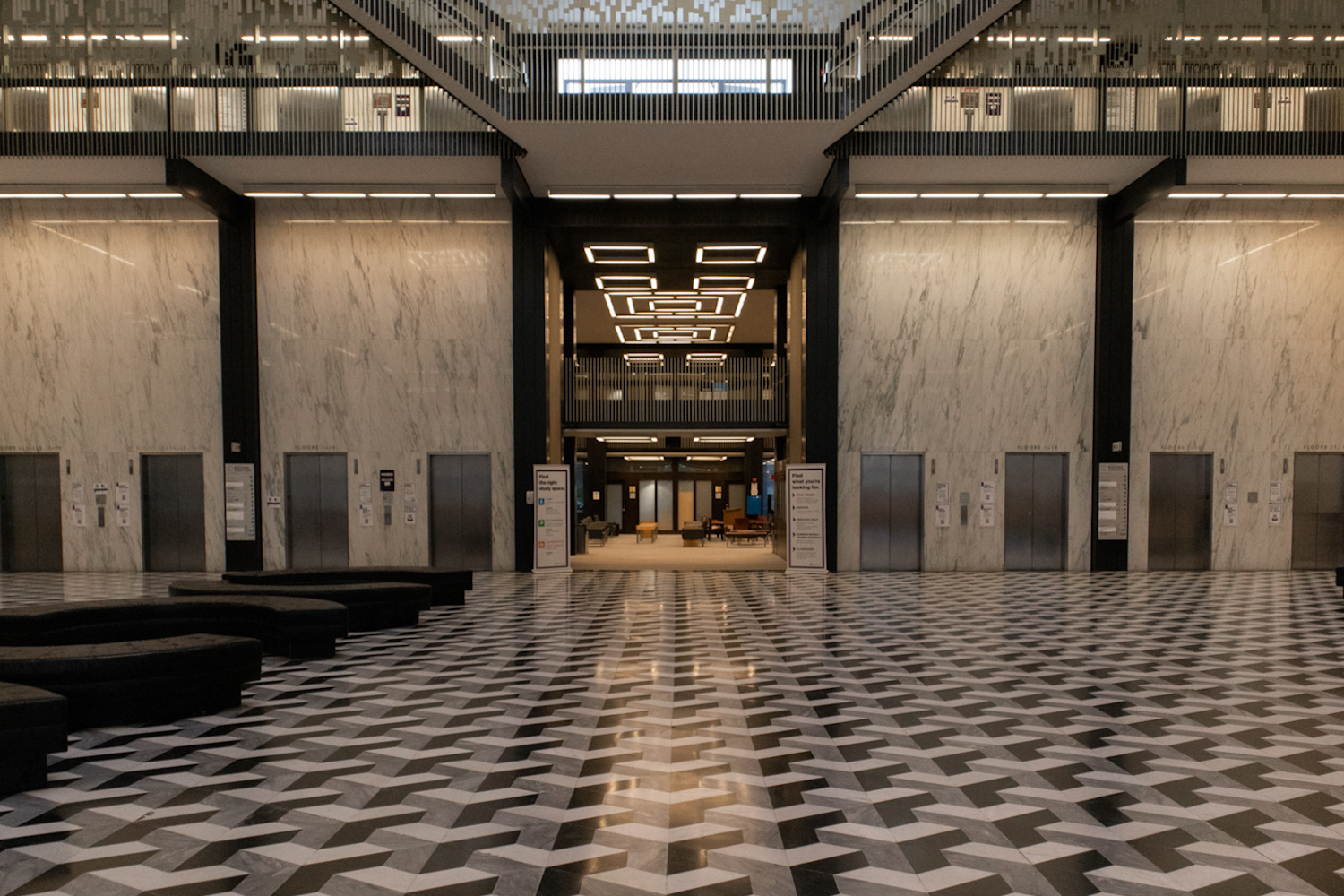 A photo of the empty Bobst Library lobby with the entrance to the study area in the center, a graphic ceramic floor, several black leather bench couches on the left, and eight elevators against the marble walls.