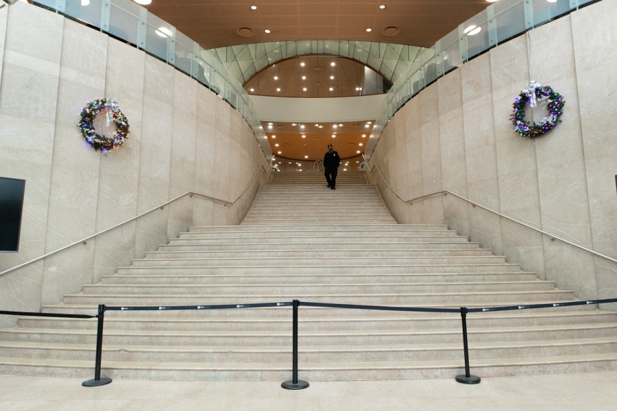 A photo of a man wearing a security uniform, walking down the lobby stairs of the Kimmel Center for University Life. Two Christmas wreaths hang on the walls symmetrically next to the stairs.