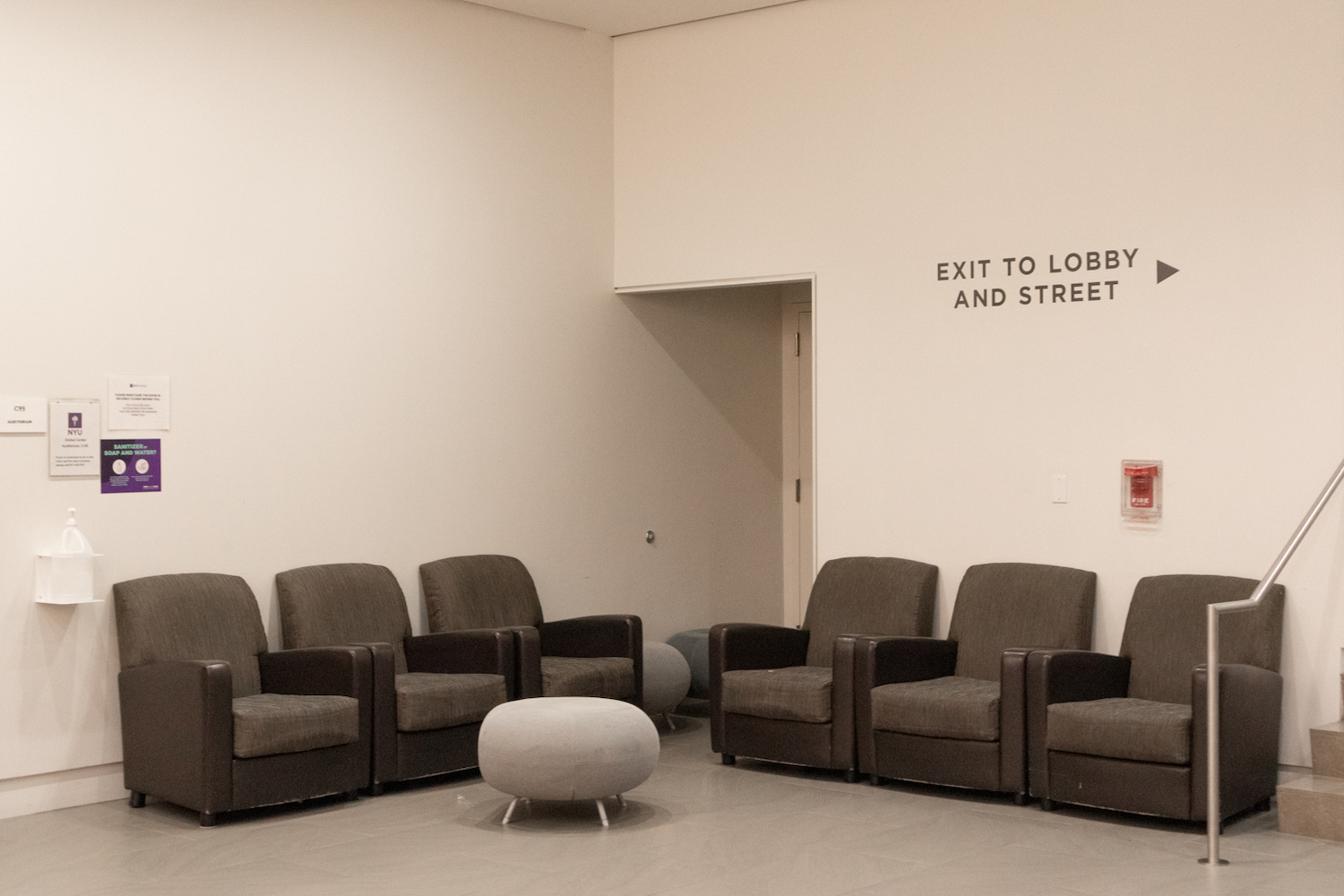 A photo of a vacant corner with six unoccupied gray couches with a sign that reads “Exit To Lobby And Street” on the right wall.