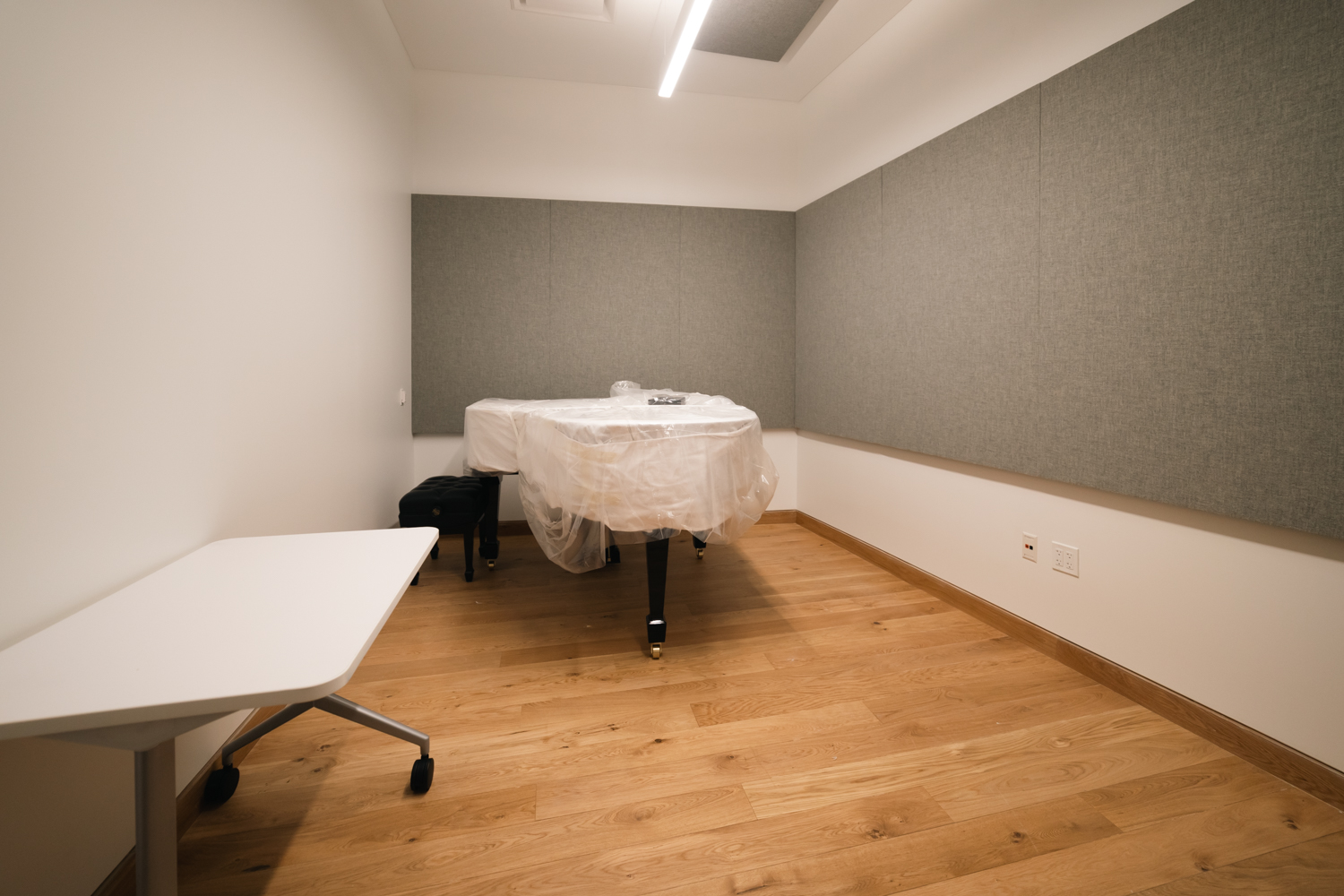 A small room with a grand piano wrapped in white paper placed against a wall. On the wall are gray acoustic panels made of fabric.