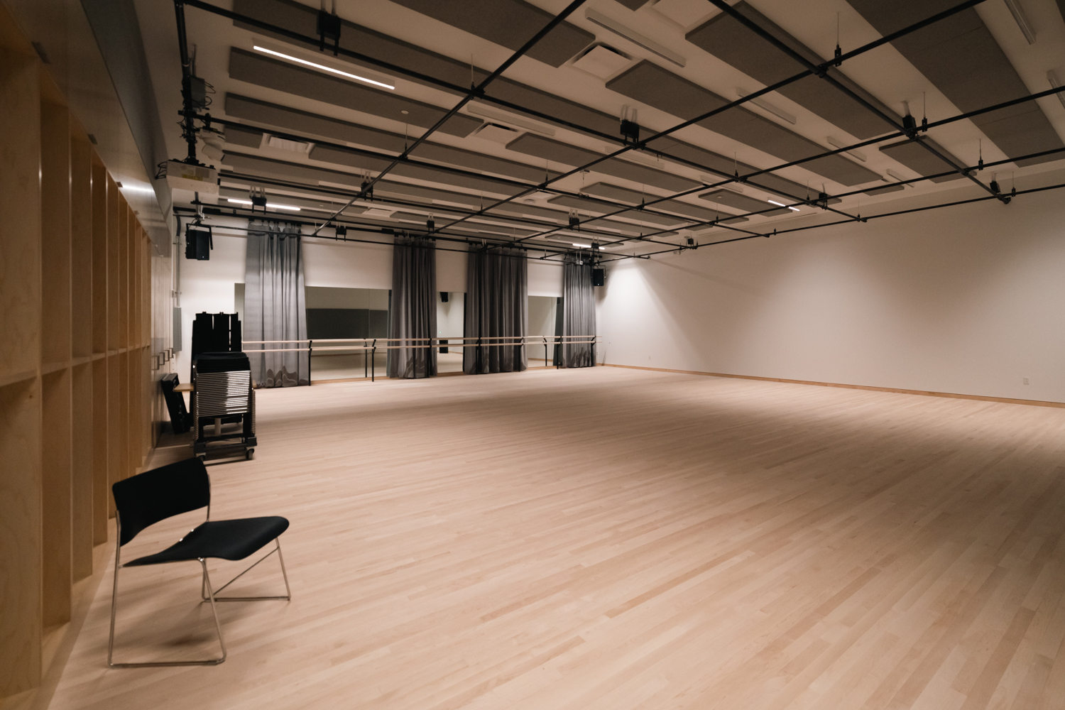 An empty rehearsal room with wooden floors and a wall of floor to ceiling mirrors. There are gray curtains and a stack of chairs in front of the mirrors.