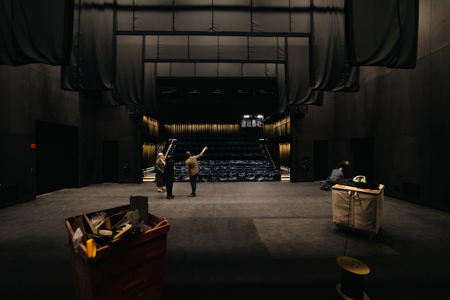A photo from the back of the stage inside a theater to an empty audience. There are four layers of black curtains hung above the stage and three people standing on the stage. Two carts of construction materials are behind them.