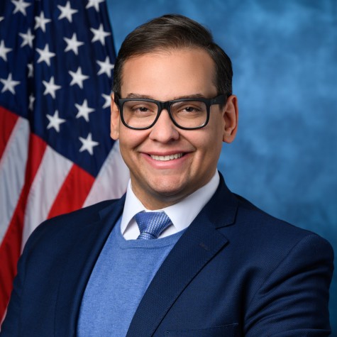 Headshot of a middle-aged man wearing black framed glasses, a navy blazer, and a blue vest, smiling in front of an American flag.