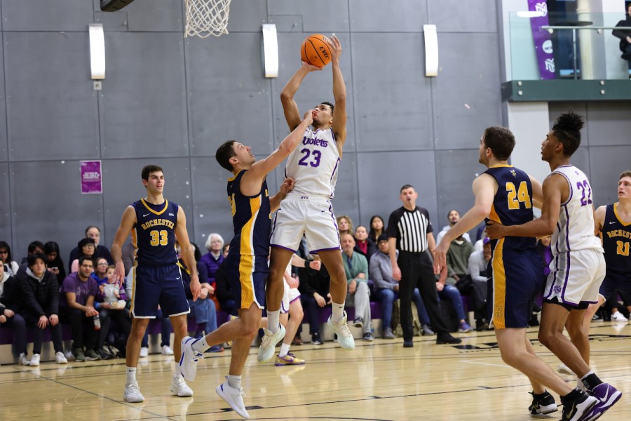 A+man+in+a+white+and+purple+jersey+and+shorts+jumping+up+toward+a+basket+with+a+basketball+in+hand.+Another+player+in+a+navy+blue+and+gold+jersey+and+shorts+puts+up+his+arm+in+an+attempt+to+deflect+him.+Other+players+in+similar+jerseys+surround+them+on+the+basketball+court+as+they+play+in+front+of+a+crowd.