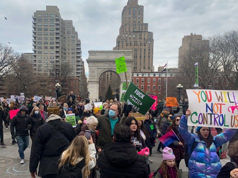 A large group of people gathers in front of the arch in Washington Square Park, wearing winter coats and holding signs. One brightly colored sign reads “you will not control us.”