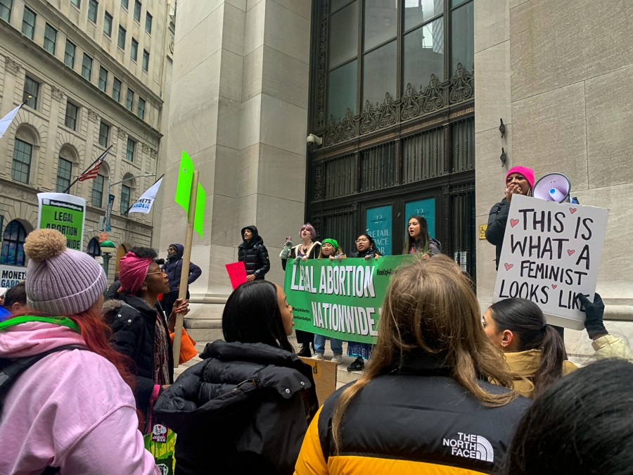 Four people hold a large, green banner reading “legal abortion nationwide” in front of a white building. A group is gathered in front of the banner holding signs. One protester shouts into a megaphone and another holds a white poster board reading “this is what a feminist looks like” decorated with pink hearts.