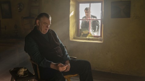 A man with a sad expression, sitting in a dimly lit room, smoking, with a window in the background. There is another man outside, looking into the room with a sad expression on his face.