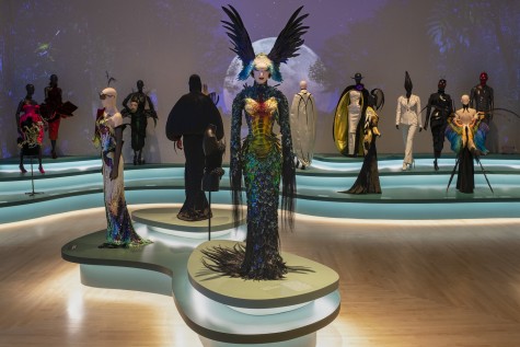 Fifteen visible haute couture dresses, gowns, coats stand in a showroom with a background illustrating a forest at night with a full moon in the sky. The centerpiece is Mugler’s “ Le Chimère,” a set of dress and headwear with colorful, beetle-like scales and dark feathered wings.