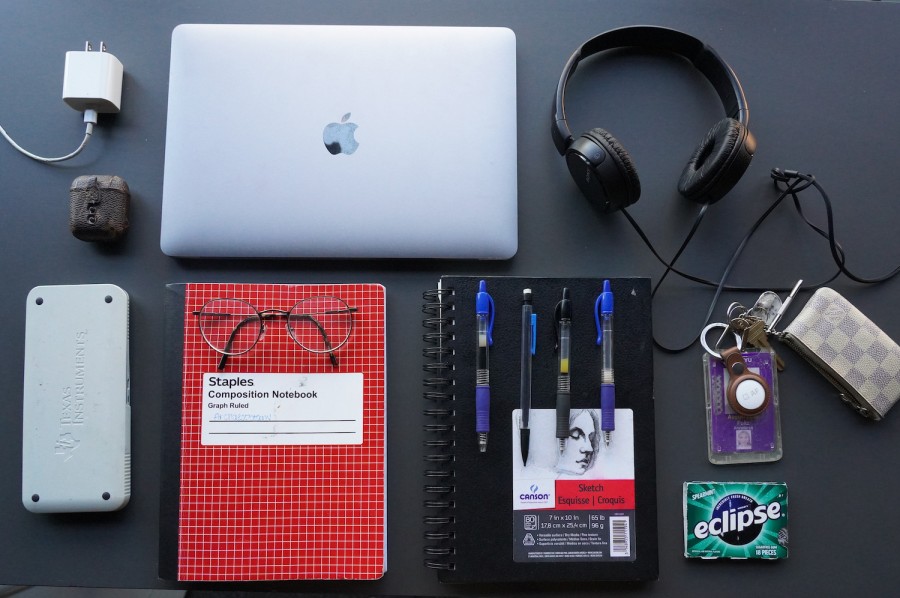 Several items laid across a gray table top, including a gray laptop, a white phone charger, a leather case for Airpods, a light gray calculator with a case, a red notebook, a pair of glasses, a black notebook, four pens, a pack of chewing gum, several keys with an N.Y.U I.D. card, a small white purse, and a pair of black headphones.