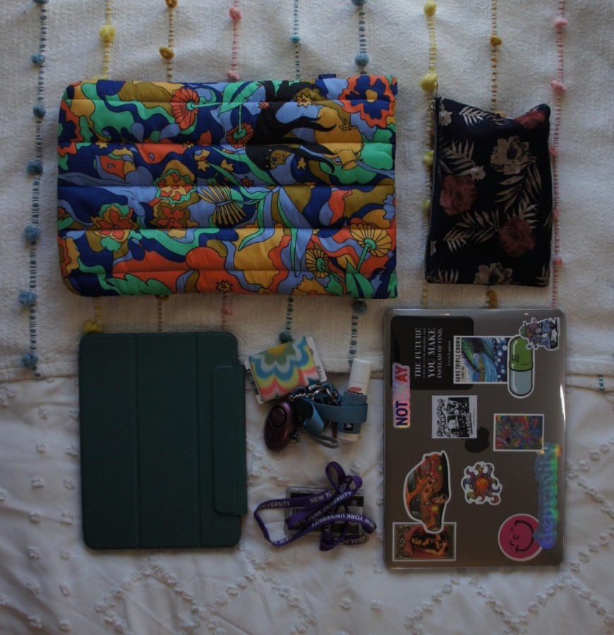 Several items laid out on a white sheet, including a laptop sleeve decorated with floral patterns, an iPad in a green case, a wallet with floral patterns, a gray laptop with many stickers, an NYU ID card attached to a lanyard, and another lanyard with a tube of lipstick.