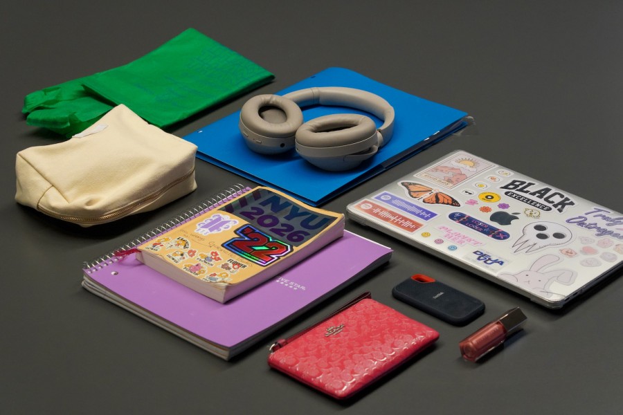 Several items laid across a gray table top including a green bag, a light yellow bag, a blue plastic folder, a pair of gray headphones, a purple notebook, a yellow notebook with an “N.Y.U. twenty-twenty six” sticker, a red handbag, a tube of red lip gloss, a portable hard drive, and a silver laptop with stickers.