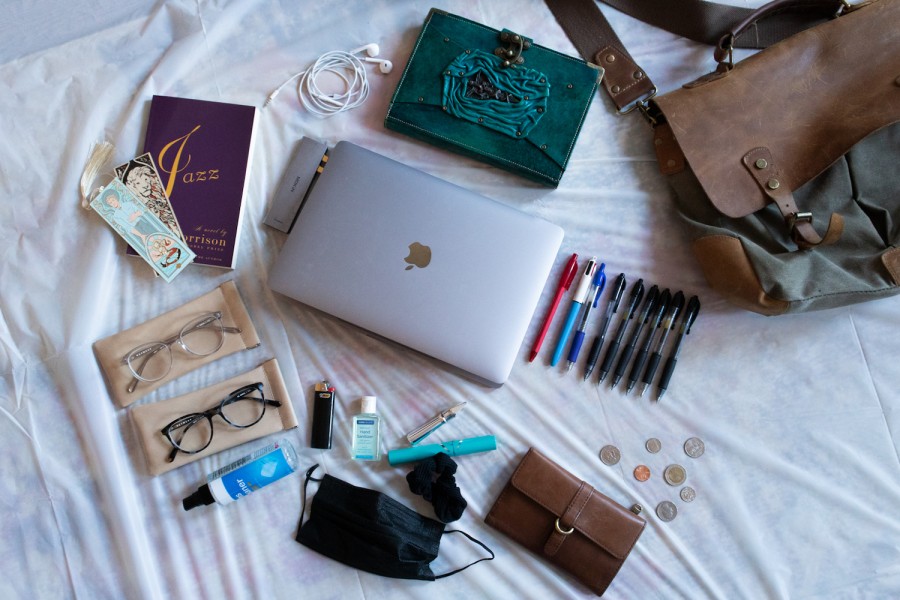 Several items strewn across a white sheet including a green journal with a leather cover, a gray laptop, a book titled “Jazz” by Toni Morrison, two bookmarks, a pair of white earphones, two pairs of glasses, a small bottle of hand sanitizer, several coins, nine pens, a container of lip gloss, a brown leather wallet, and a brown leather tote bag.