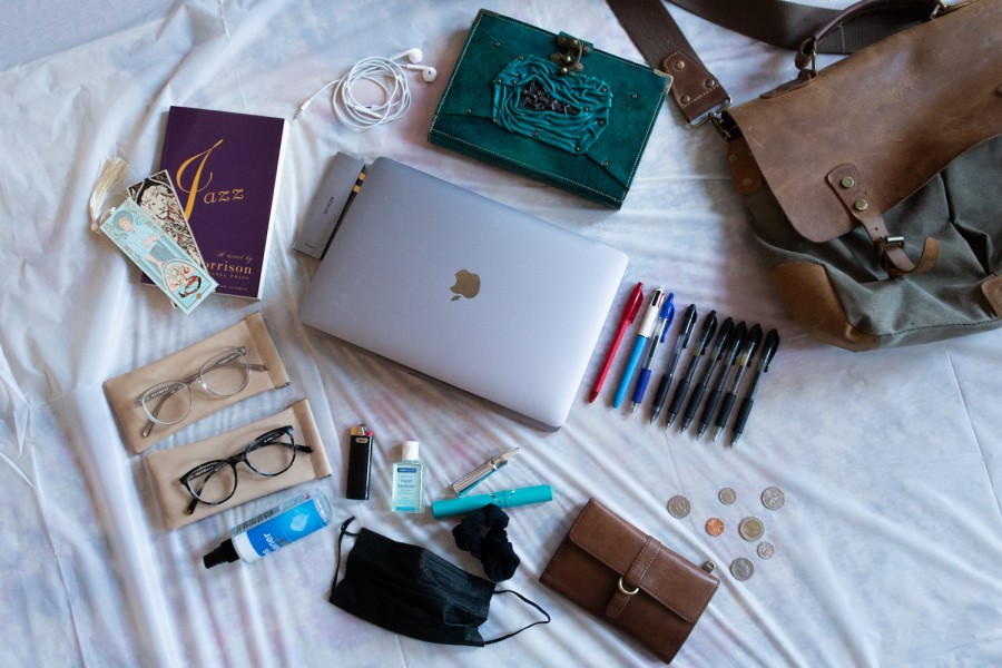 Several items are scattered on the white sheet, including a green leather-bound magazine, a gray laptop, Toni Morrison's Jazz book, two bookmarks, a pair of white headphones, two pairs of glasses, a bottle of hand sanitizer.  , some coins, nine pens, a jar of lip gloss, a brown leather wallet, and a brown leather tote bag.