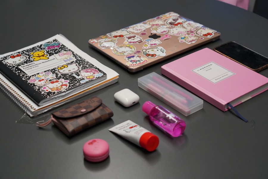 Several items laid across a gray tabletop including a notebook with Hello Kitty stickers, a pink laptop with Hello Kitty stickers, a smartphone, a pink notebook, a plastic pencil box with three pink pens inside, a pink bottle of hand sanitizer, a Hello Kitty themed hand cream and lip balm set, a pair of wireless headphones, and a Coach wallet.