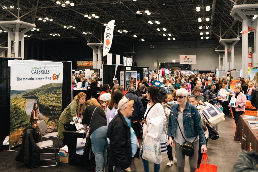 A large group of people walking through a crowded convention center that has travel-related booths on the side.