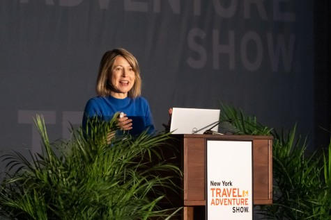 A woman with short, blonde hair and a blue sweater making a speech behind a podium with the text “New York Travel Adventure Show.