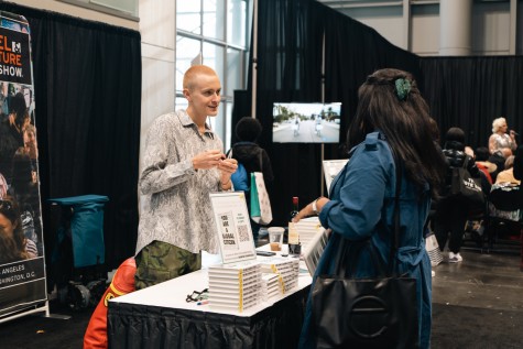 A man with short hair and a patterned, white shirt talking to a woman wearing a blue coat. In between them is a book signing table with several stacks of books and a small sign reading “You are a global citizen.”