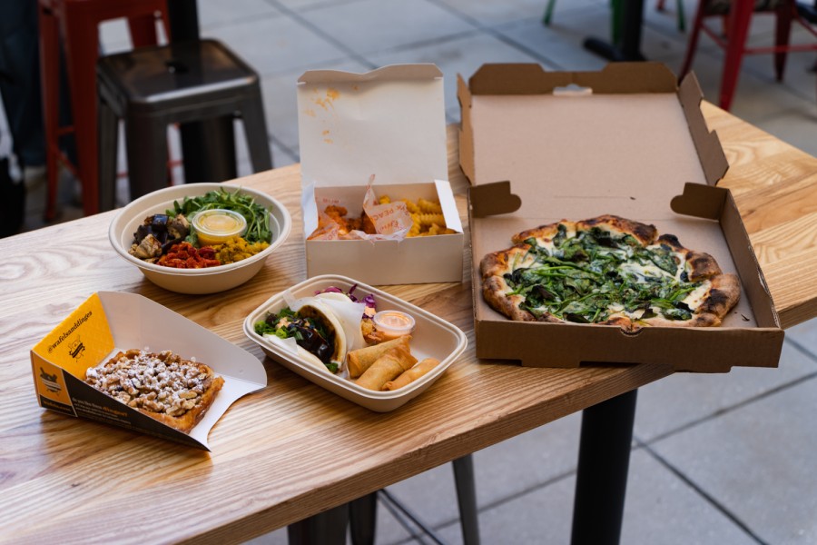 Five containers of food placed on a wooden table. From the left to right: a waffle with walnuts in a yellow paper container; two bao buns and three egg rolls in a paper box; a salad bowl with brown rice, eggplant, arugula, tomatoes and curried chickpeas; a box of fried chicken and fries; and an entire pizza with goat cheese and spinach in a pizza box.