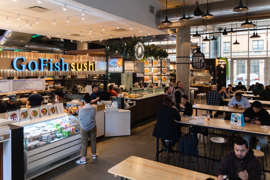 The interior of a food court with a sushi counter and several people eating at rows of tables next to the counter. In the back are floor-to-ceiling windows.