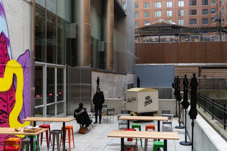 A patio outside of a food court with two people sitting at a black, round table. Four wooden tables and several high stools of different colors are on the ground. There is a mural on the wall next to a glass entrance.