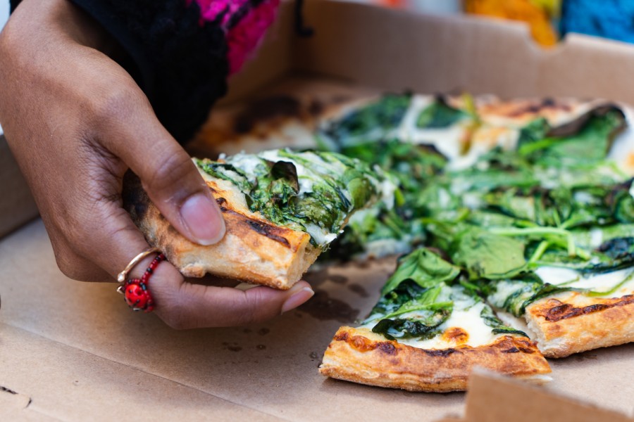 A hand with two rings on the index finger holds a slice of goat cheese and spinach pizza in a pizza box.
