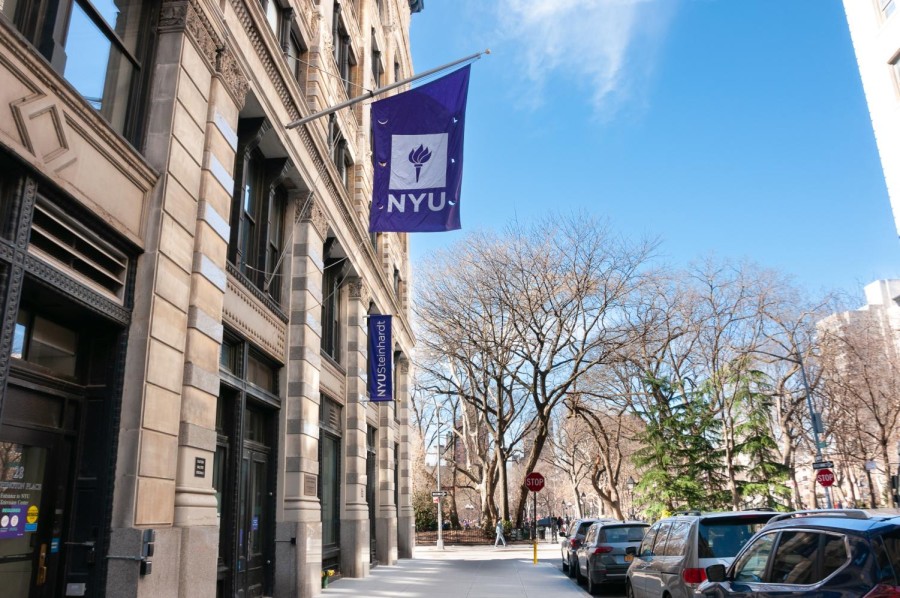 An N.Y.U. flag hangs above the Steinhardt School of Culture, Education, and Human Development. Cars line the road to the right of the building and an entrance to Washington Square Park can be seen in the background.