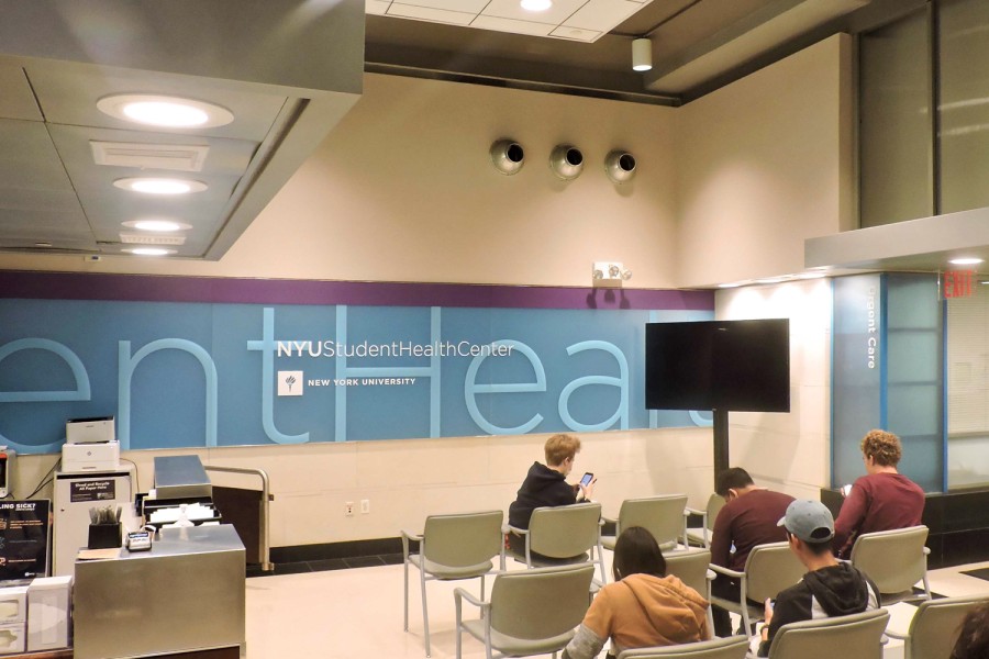 The waiting area of the NYU Student Health Center. A man wearing a black hoodie is on his phone, sitting in a row of chairs closest to the front of the room. Two men with maroon shirts sit in the second row, and another two students sit in the third row. A reception desk is on the right side of the room.