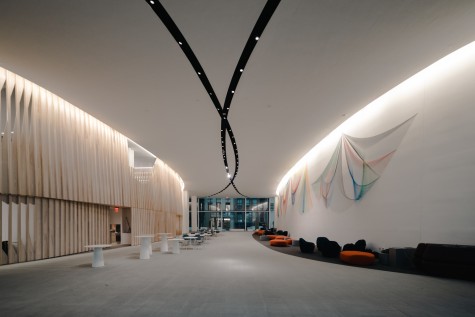 A large, open lobby with two stripes of lights on the ceiling. There are white tables on the left side and red sofa chairs on the right side. An art installation with strands of colorful wires hangs on the wall on the right.