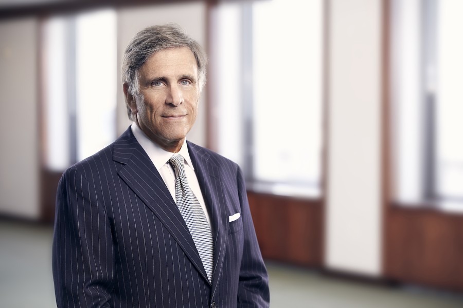 A man in a dark blue striped suit with a checkered tie facing forward with his arms crossed. Behind him is a wall with multiple tall windows.