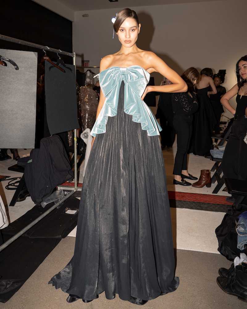 A full body flash photograph of a model standing backstage, wearing a light blue, velvet, bow-shaped top and long, pleated, black velvet dress.