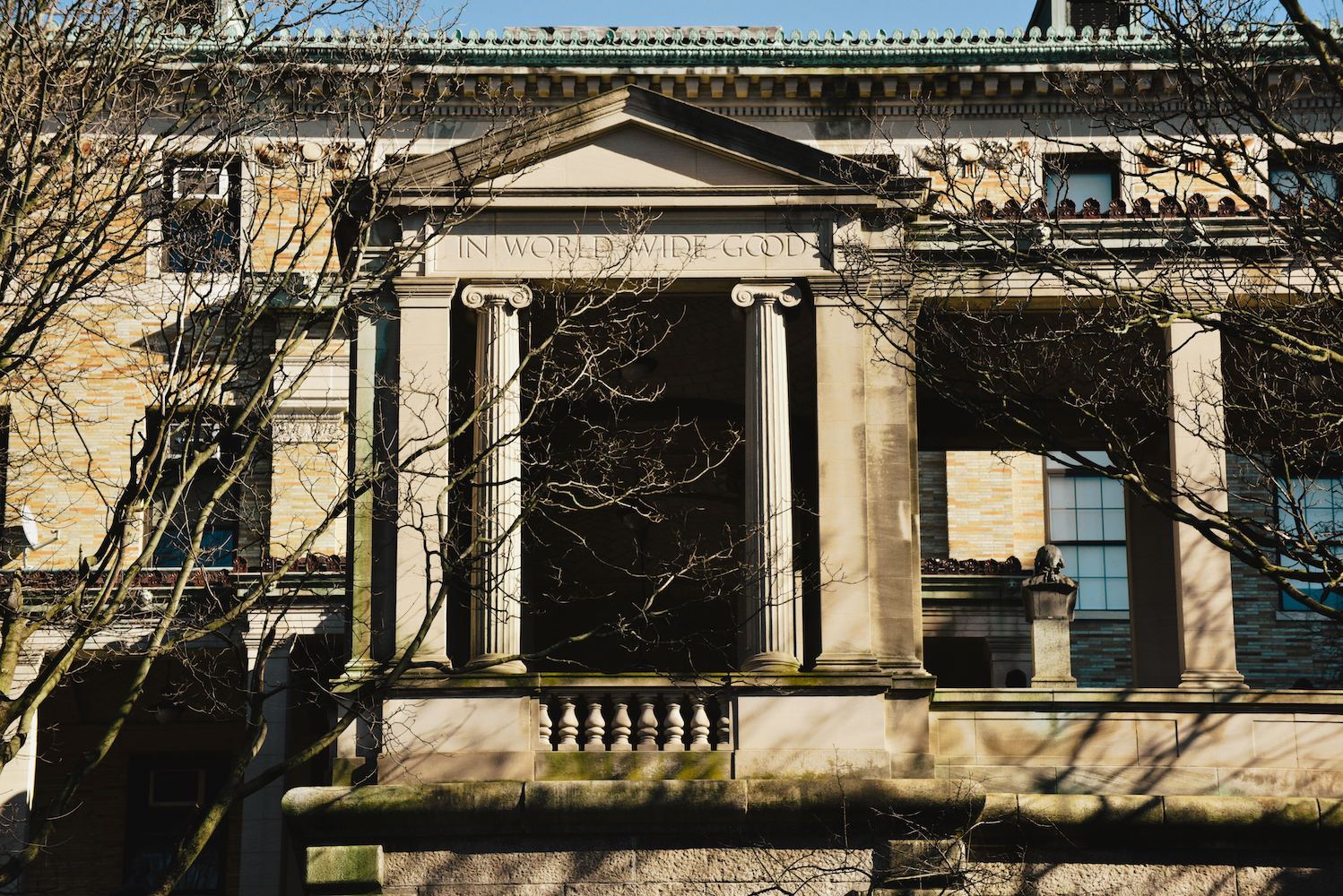 An greek style entrance of a building with marble façade. Below the pediment is an inscription that reads "In World Wide Good."