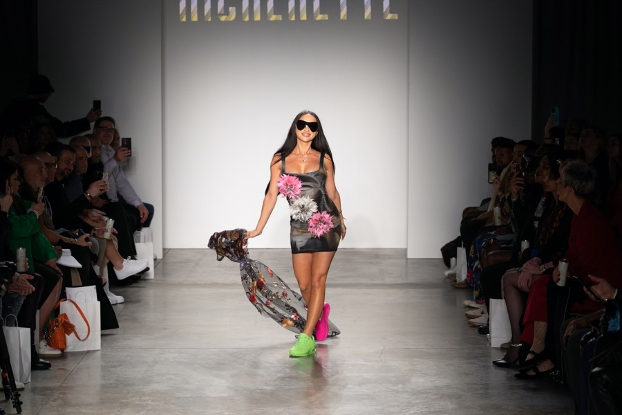A female model walking down the runway wearing a black dress decorated with white and pink flowers, and a pair of green and red sneakers.