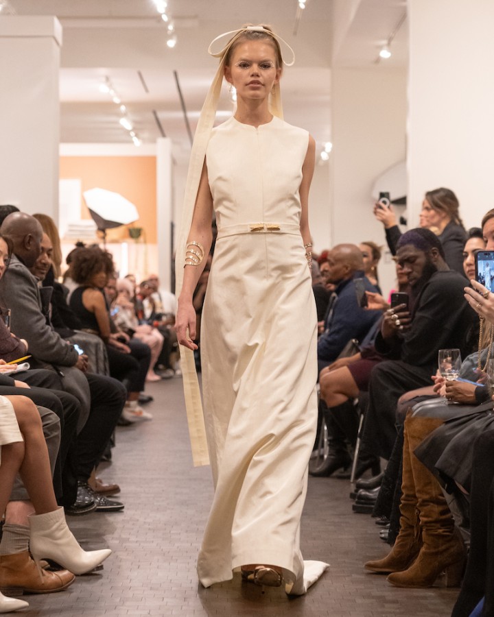 A model walking down the runway wearing a white silk dress with gold accent jewelry, a long tan ribbon in her hair, gold heels and pearls decorating her eyelids.