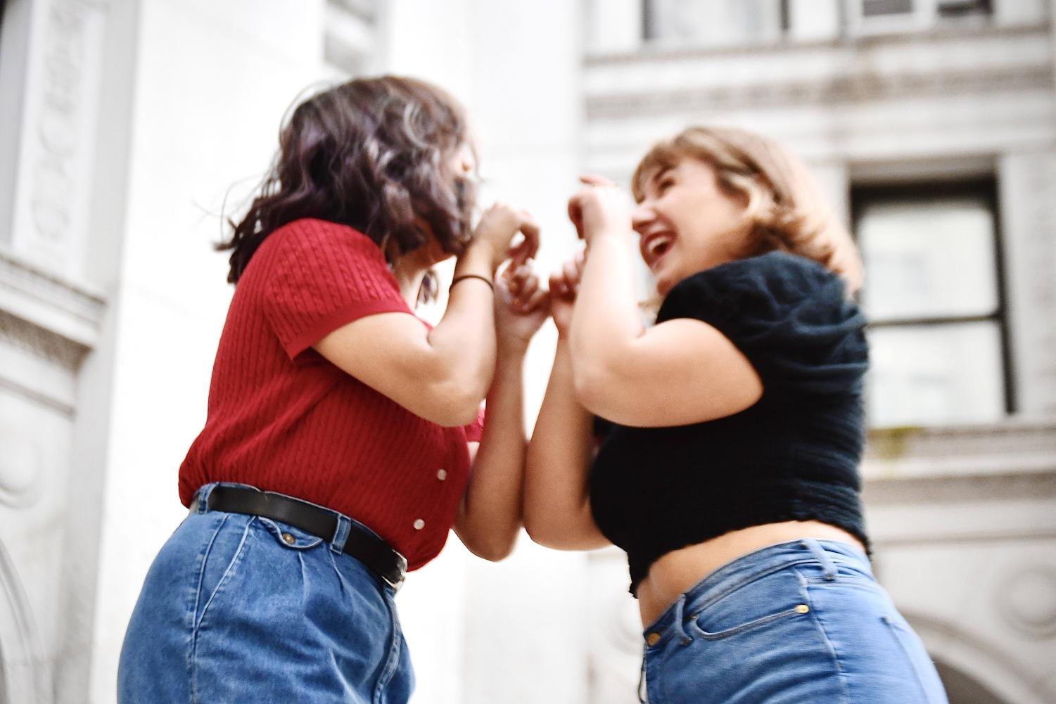 Two people jump toward each other excitedly. One has blonde, short hair, and is smiling. They are wearing a black crop top and blue jeans. The other person, on the left, wears a red short-sleeved sweater and blue jeans.