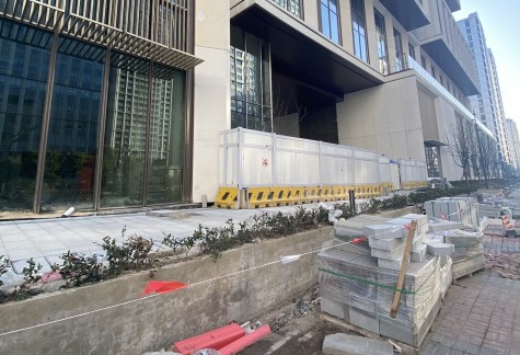 The exterior of a modern glass building still under construction. The bricks to be laid on the sidewalk are stacked on the ground.