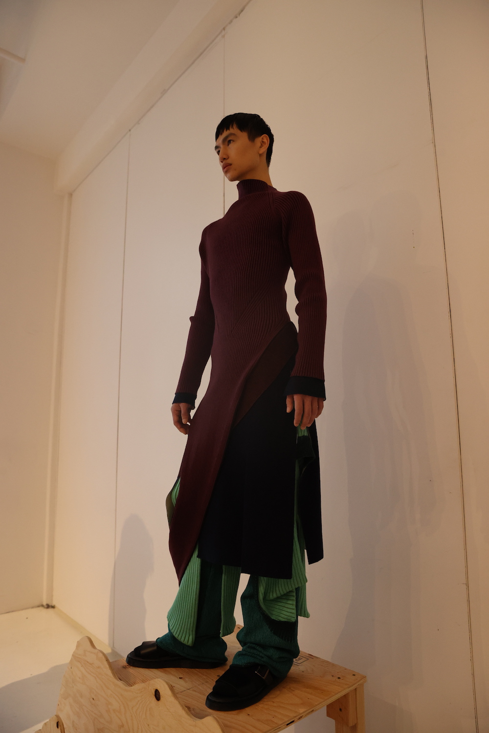 A model wearing a burgundy robe, green knitted pants and black sandals.
